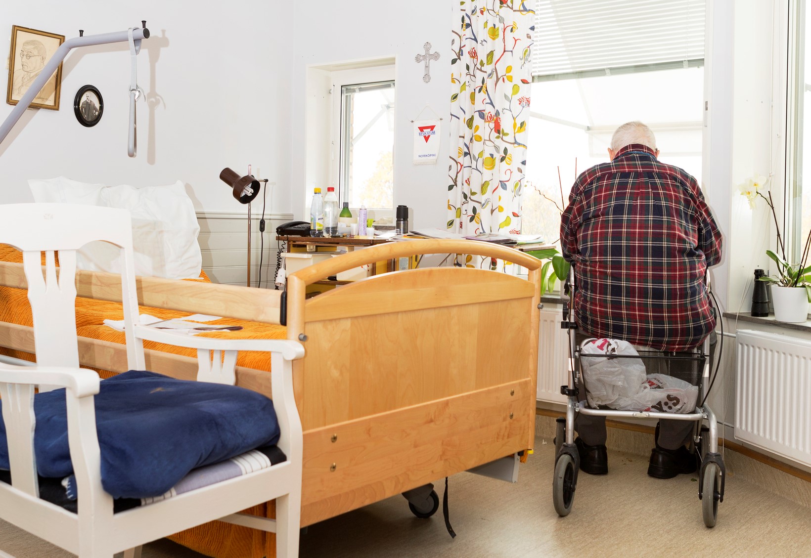 En äldre person i en vårdmiljö som sitter på sin rollator och tittar ut genom ett fönster.