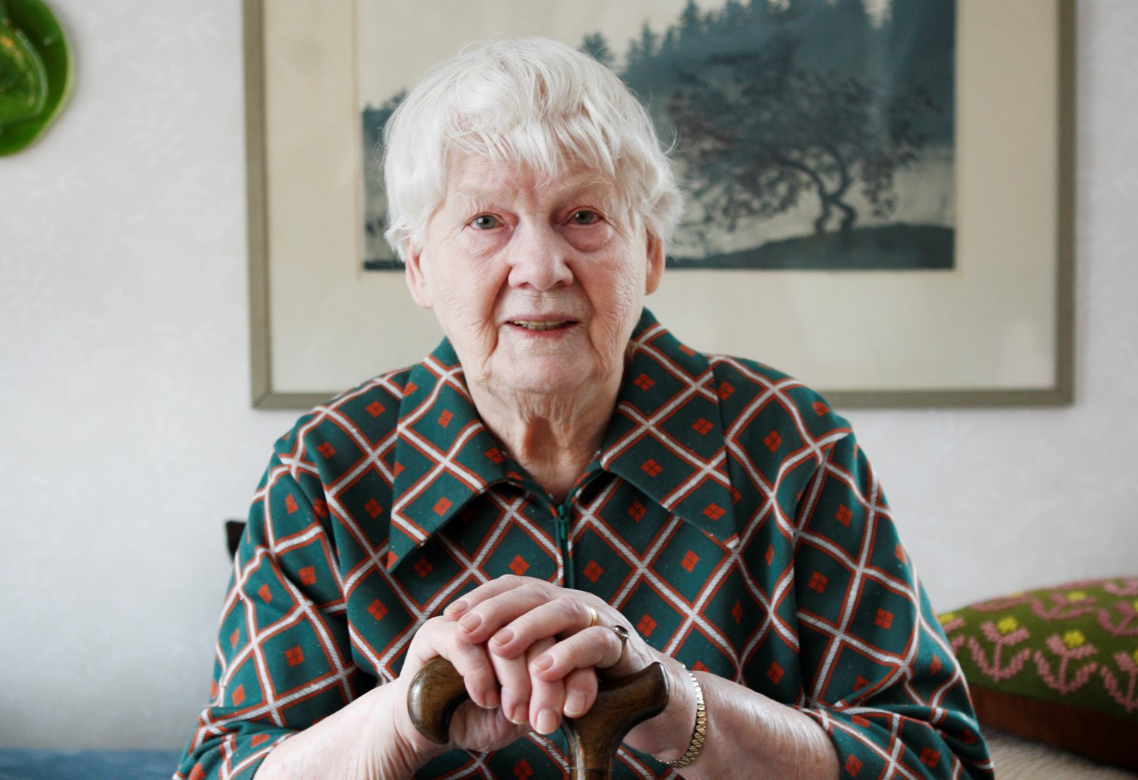 Äldre kvinna sitter ned och har händerna på en käpp