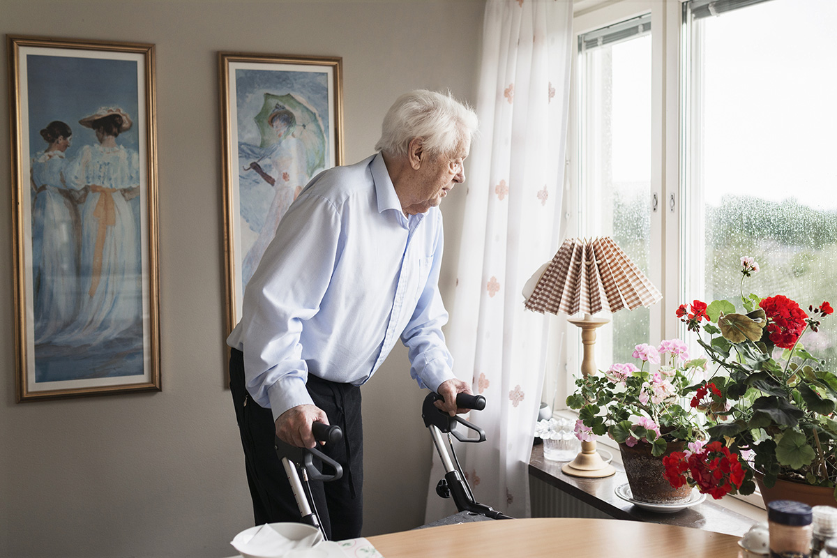 Äldre man stödjer sig på sin rollator och tittar ut genom fönstret